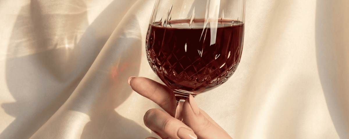 A hand holds a wine glass with red wine against a silky fabric backdrop.